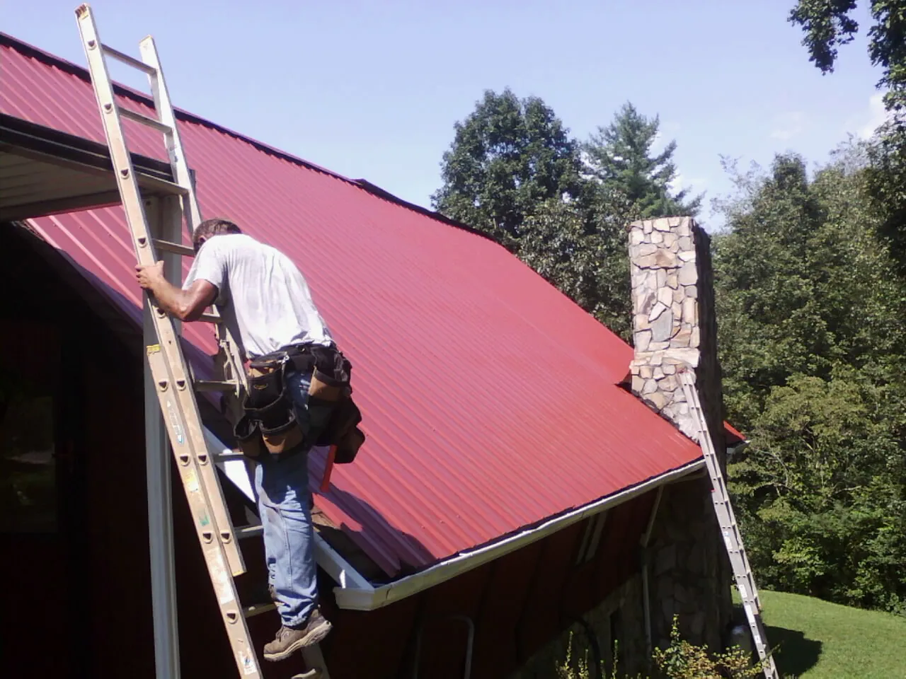 Roofer on a ladder beside a red metal roof and stone chimney — Soffit & Fascia Repair by Roofing Brothers in Fairless Hills